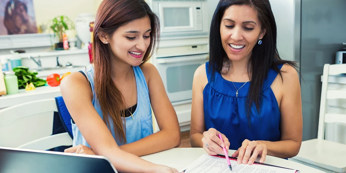 A parent and a student sit at a table in the kitchen looking and writing on a paper